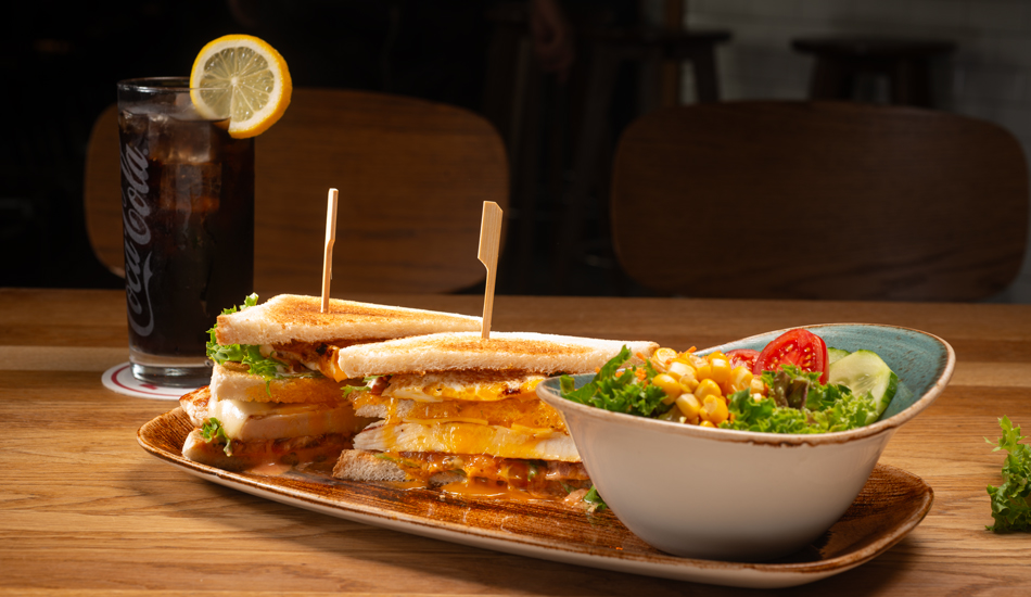 Das Salversandwich mit Toast, Hähnchenbrust, Speck, Salat, Ei, Tomate, Käse, Mayonaise, Cocktail-Dip, Dazu Beilage nach Wahl. Dahinter steht eine Coca Cola, daneben eine kleine Salatschüssel.