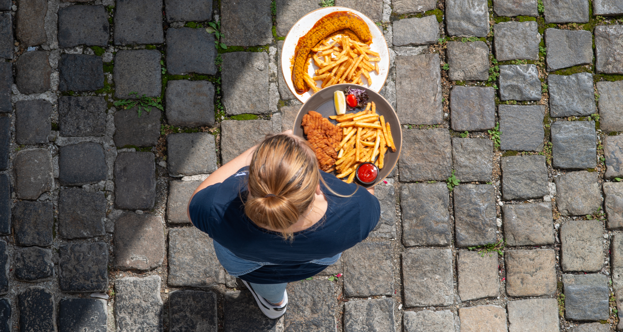 Eine Mitarbeiter aus der Vogelperspektive fotografiert, trägt gerade zwei Teller mit Currywurst und Schnitzel zu den Gästen. Zusehen ist der typische Boden aus Kopfsteinpflastern in Pfaffenhofen.